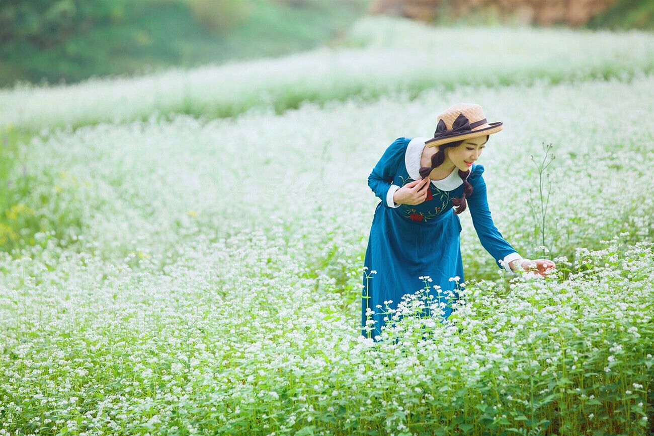 A person in a blue dress and hat walking through a field of white flowers.