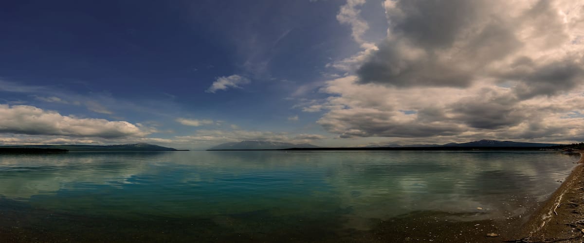 A calm lake with clear blue water reflecting the sky, surrounded by mountains and a cloudy sky.