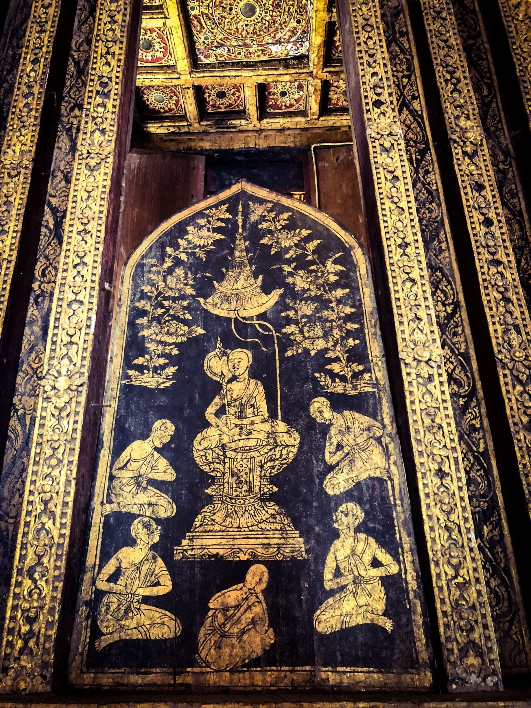 A detailed mural with a central figure seated on a lotus pedestal, surrounded by smaller figures in a dark background with golden patterns.