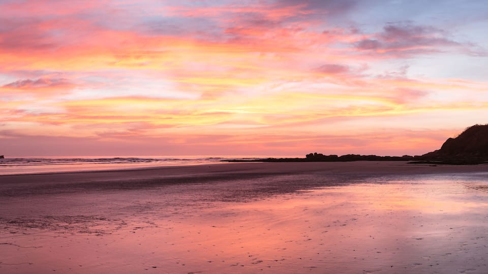 A beach at sunset with a pink and orange sky, calm water, and rocky formations in the distance.