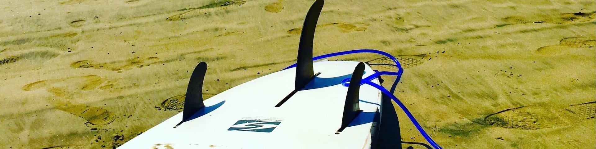A white surfboard with the number 12-5 on it, lying on a sandy beach with footprints and a clear blue sky with clouds in the background.