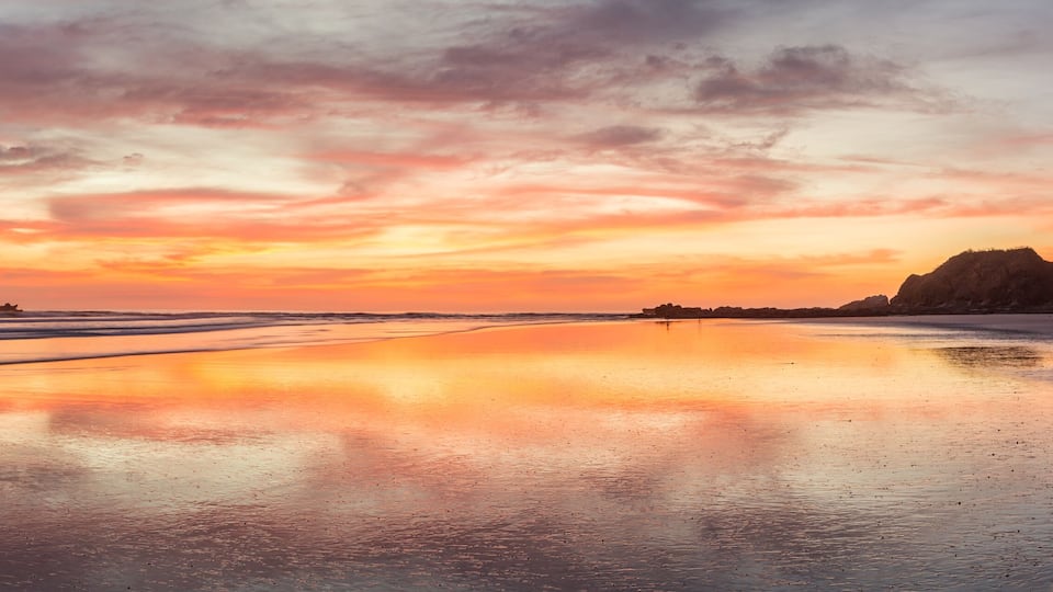 A beach at sunset with a calm sea, a sandy shore, and distant hills.