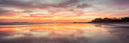 Ein Strand bei Sonnenuntergang mit ruhiger See, sandigem Ufer und weit entfernten Hügeln.