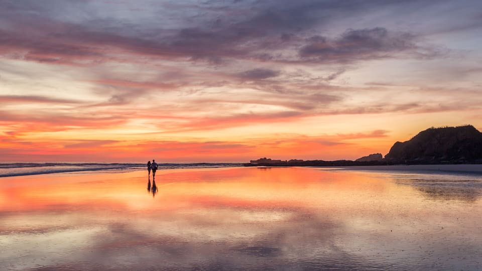 A beach at sunset with a calm sea, two people walking, and a distant rocky outcrop.
