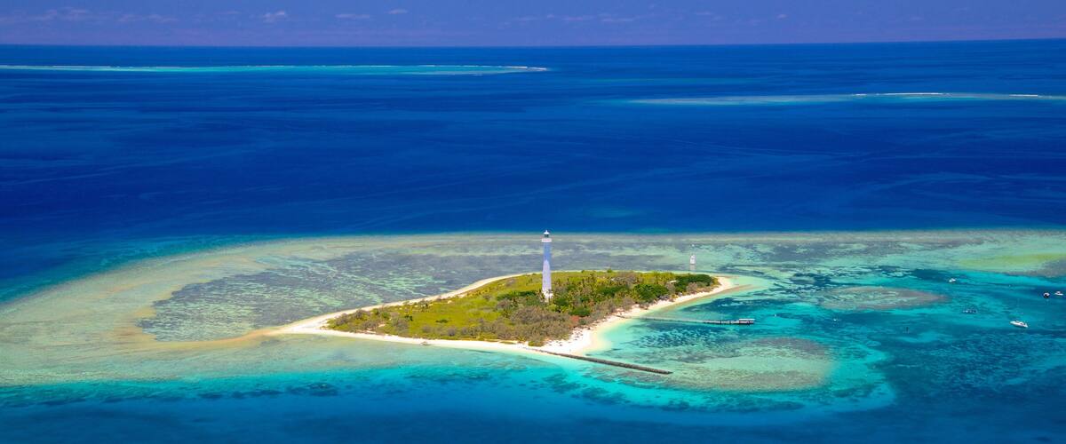 An aerial view of a turquoise island with a lighthouse, surrounded by clear blue waters and coral reefs.
