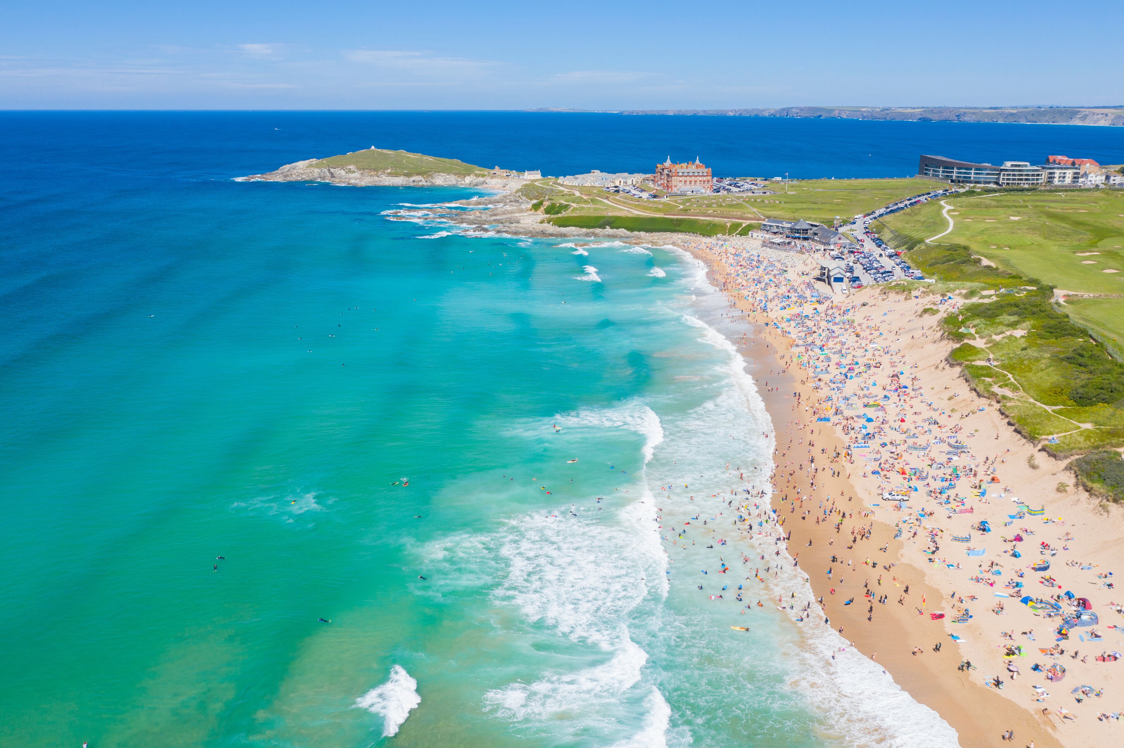 A coastal beach with clear turquoise waters, a sandy shore, and a large building complex in the background.