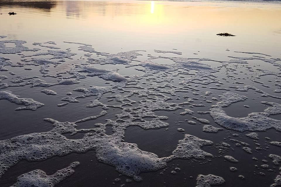 A beach with foam patterns on the sand and a sunset in the background.