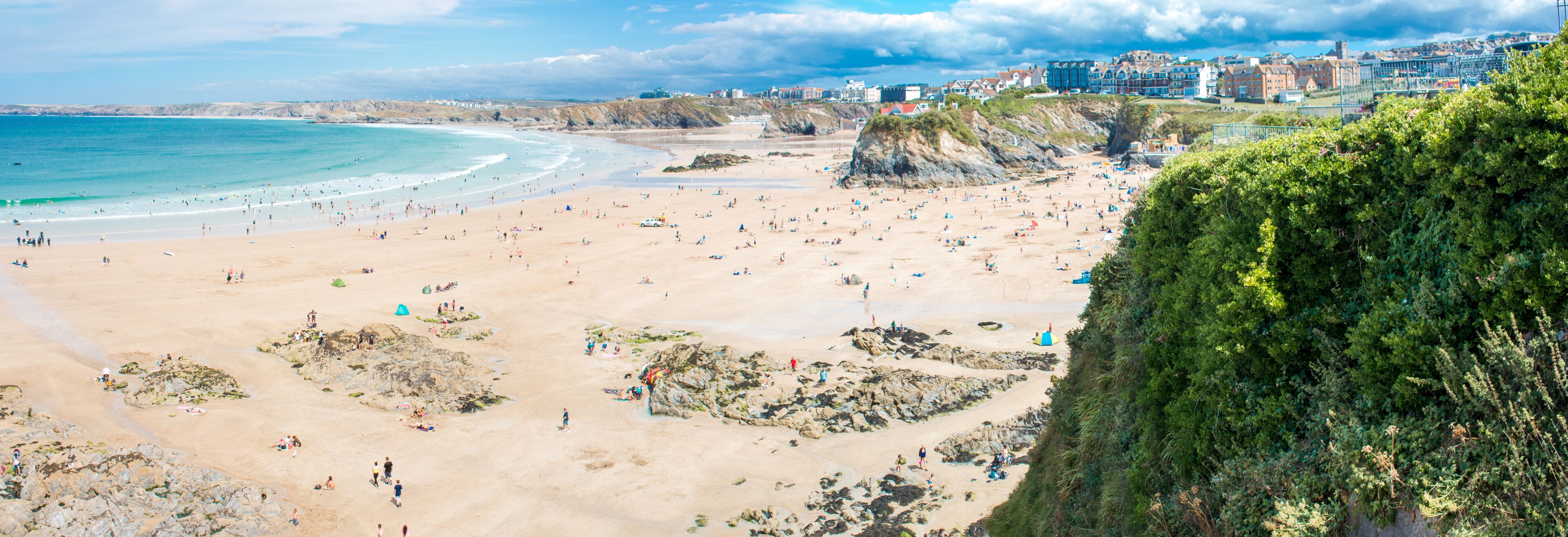 A sandy beach with rocky outcrops, a coastal town in the background, and a cliff with greenery.