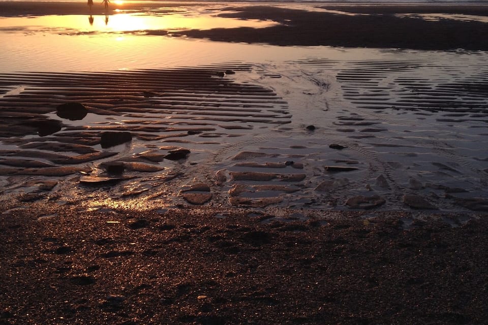 Praia ao pôr do sol, com pegadas e areia molhada.