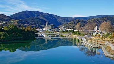 A serene lake reflecting a town with a prominent clock tower, surrounded by lush green hills and mountains.