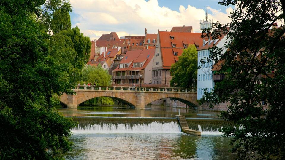 A stone bridge over a river with a fountain, surrounded by lush greenery and historic buildings with red-tiled roofs.