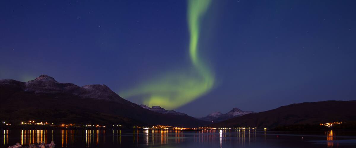 A night scene with the Northern Lights over a calm lake, mountains in the background, and a lit shoreline.