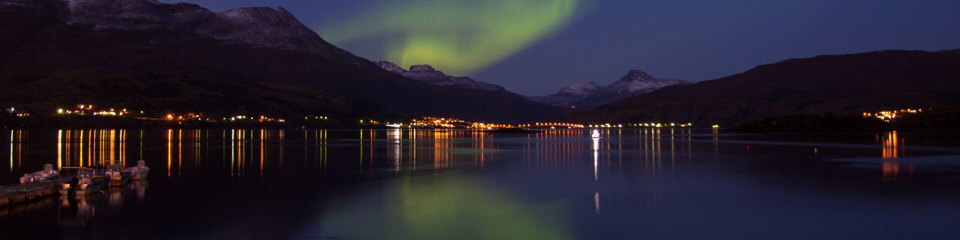 A night scene with the Northern Lights over a calm lake, mountains in the background, and a lit shoreline.