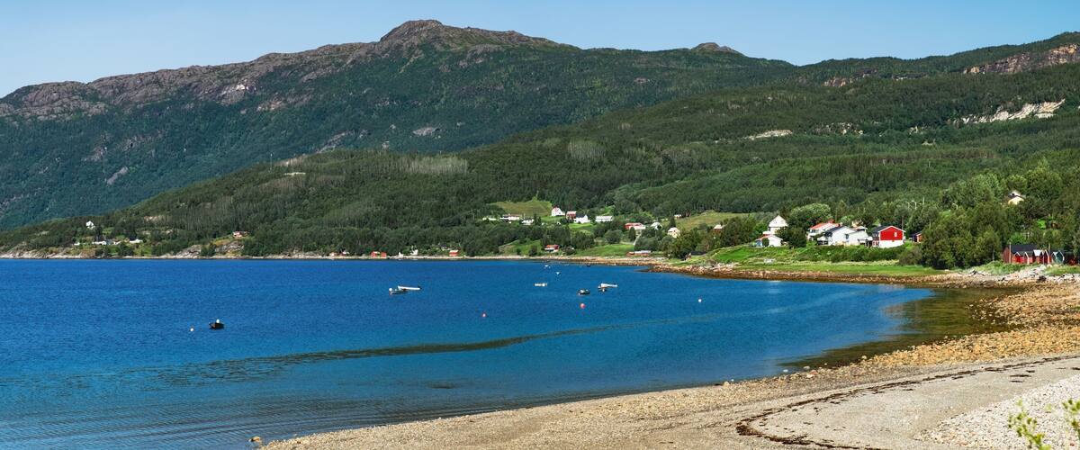 A coastal landscape with a clear blue lake, a rocky shoreline, and a backdrop of green hills with scattered houses.
