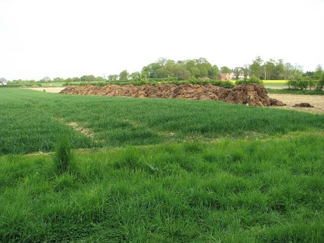 View across fields. A cereal crop can be seen growing in the field at left and the oilseed rape is in flower in the distant field seen at right. A large heap of muck has been deposited by the edge of the bare field wedged in between the two, where a crop of maize stood until recently, providing food and shelter for game birds such as pheasants and partridges over the winter. The field has now been cleared and will soon be ploughed. Whitwell Hall Farm can be seen in the distance (in adjacent grid square).