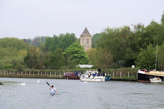 A person paddling a kayak on a river, with boats docked along the shore and a tower in the background.