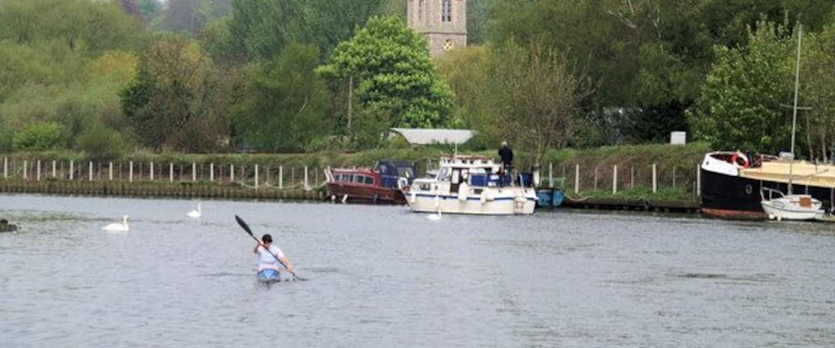 A person paddling a kayak on a river, with boats docked along the shore and a tower in the background.