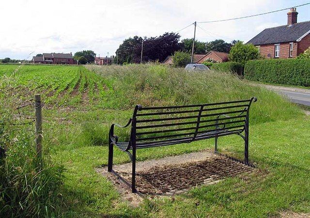A bench in a grassy field with a road and houses in the background.