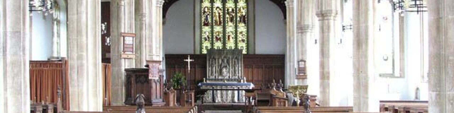 Interior de igreja com bancos de madeira, um corredor central e uma janela de vitrais.