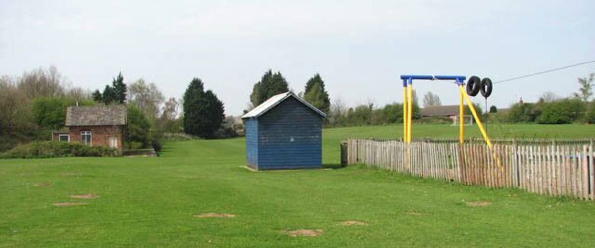 A grassy field with a blue shed, a wooden fence, and a blue swing set.