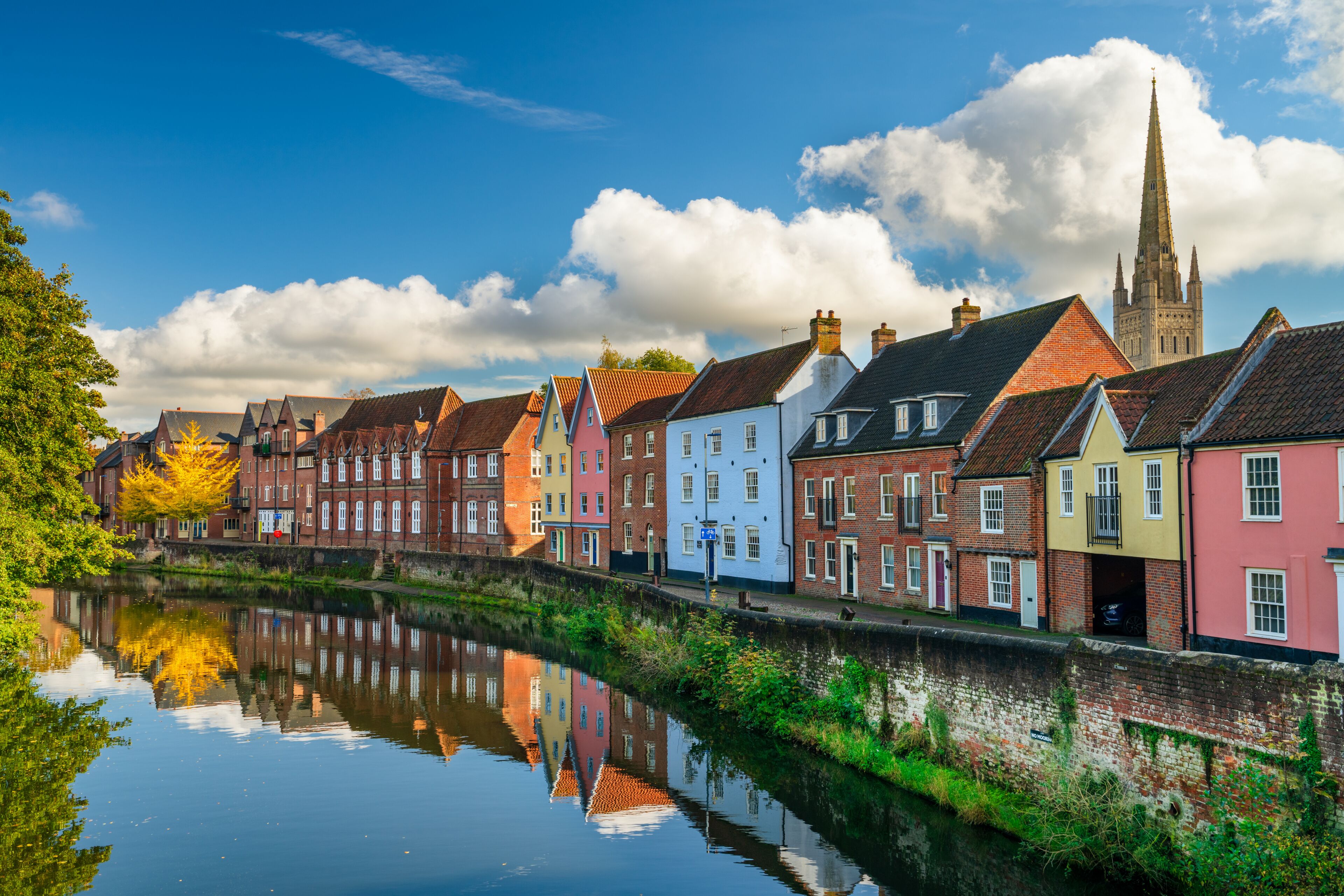 A canal with colorful houses reflecting in the water, a church spire in the background, and a clear blue sky.
