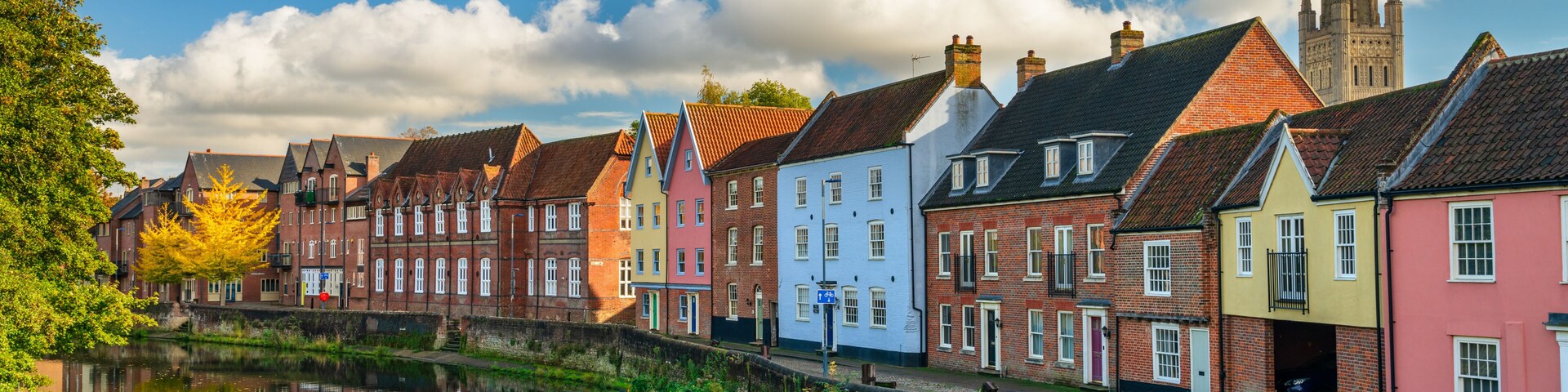 Un canal con casas coloridas reflejándose en el agua, una aguja de iglesia al fondo y un cielo azul despejado.