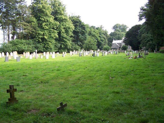 A cemetery with numerous headstones and crosses, surrounded by trees and a building in the background.