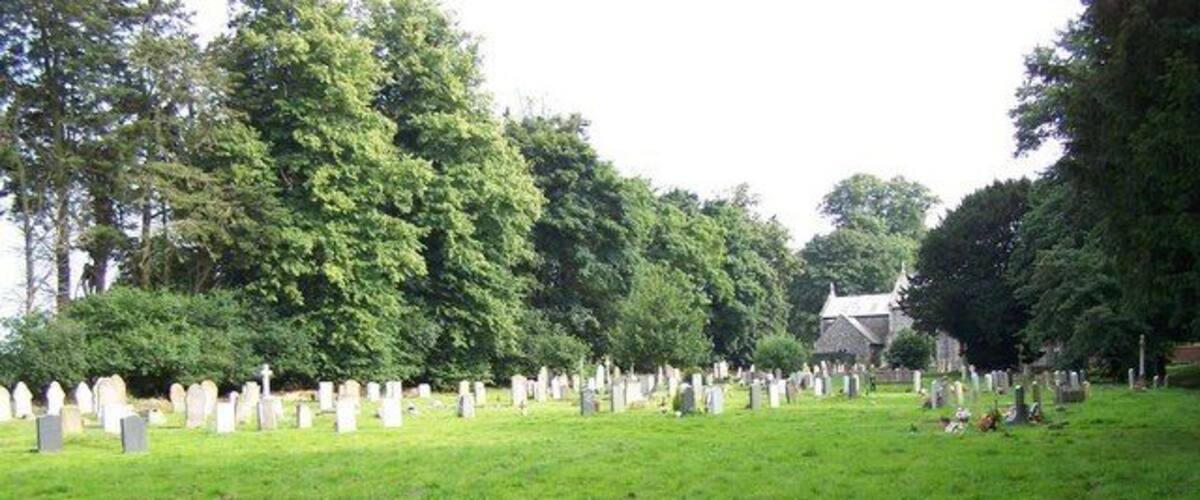 A cemetery with numerous headstones and crosses, surrounded by trees and a building in the background.