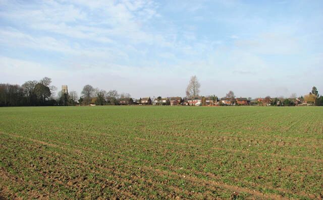 A rural landscape with a large open field, a few scattered houses, and a clear sky.
