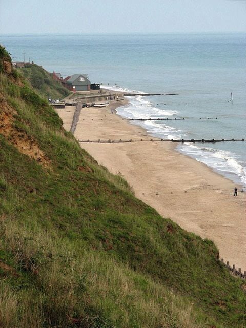 A coastal beach with a grassy hill, a wooden walkway, and a building near the water.