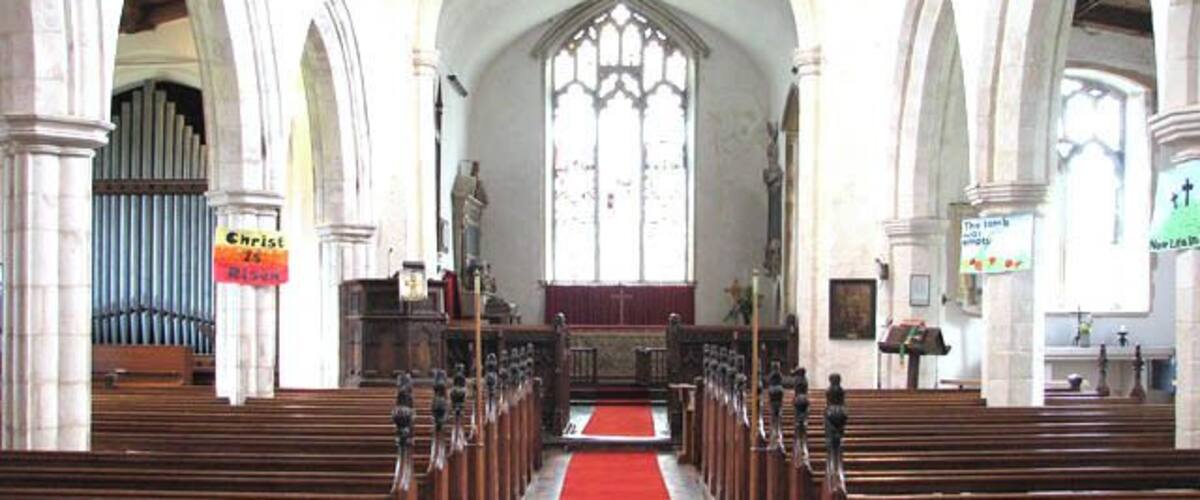 A church interior with wooden pews, a red carpet, and a large arched window.