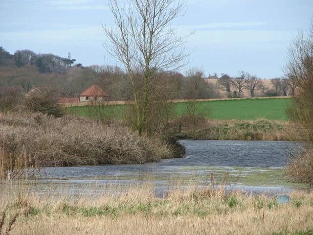 A river flowing through a rural landscape with fields, a leafless tree, and a small building in the background.