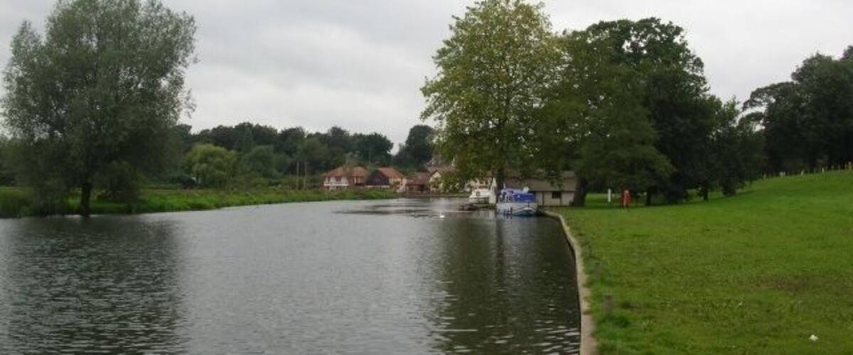 A calm river with a grassy bank, trees, and a small building in the distance.