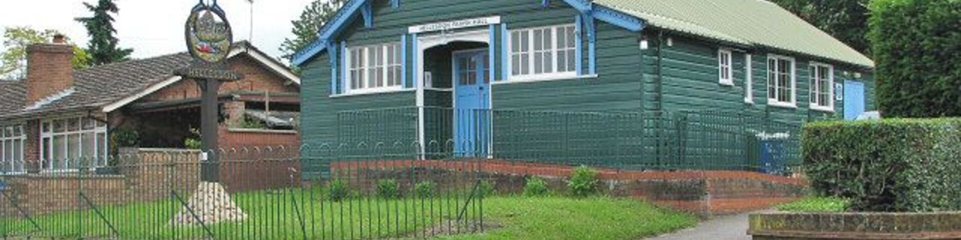 Hellesdon Parish Hall. Beside Low Road, adjoining St Mary's church; the village sign > 850755 - 850757 is located here. Hellesdon is a large suburb of Norwich. Signs of very early settlement, such as a variety of flint tools (believed to be at least 4,000 years old) have been unearthed in the area. Bronze axe heads were found near Hellesdon Hall and a skeleton dating from around 600 CE was discovered in Low Road. It is not clear where the old name for Hellesdon - Hailesduna - originates from. It is believed that Edmund, the King of the East Angles, may have been martyred here. In 985, Abbo of Fleury, who at Ramsey Abbey (Cambridgeshire) compiled the 'Life of St Edmund', writes that he heard the Archbishop relate a story that came from a young man who had heard it from a very old man who claimed to have been King Edmund's armour bearer at the time of his death. On his capture, Edmund was whipped and tied to a tree, and shot with arrows. He was then beheaded and his head thrown into a bramble thicket in Hegelisdun Wood - hence the association with Hailesduna, present-day Hellesdon. The King's head was later found, guarded by a wolf, and according to the story the body was buried in a small chapel built nearby for the purpose. The site of the chapel is believed to be the site where St Mary's church stands > 850769. The church and the dead body of King Edmund, guarded by a wolf, are depicted on the village sign > 850757.