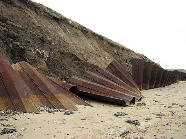 Steel sheet piling. The sheet piling, erected here behind the breakwater in an effort to protect the soft cliff face, has slumped and consequently twisted; it is also regularly being pushed over by high waves which take away whole sections of cliff. See also > 774025. A variety of sea defences have been erected along the beach north of Bacton; the defences are particularly extensive nearing the Bacton gas terminal. But despite all these measures the sea is regularly breaching the breakwaters, groynes, revetments and steel sheet pilings.