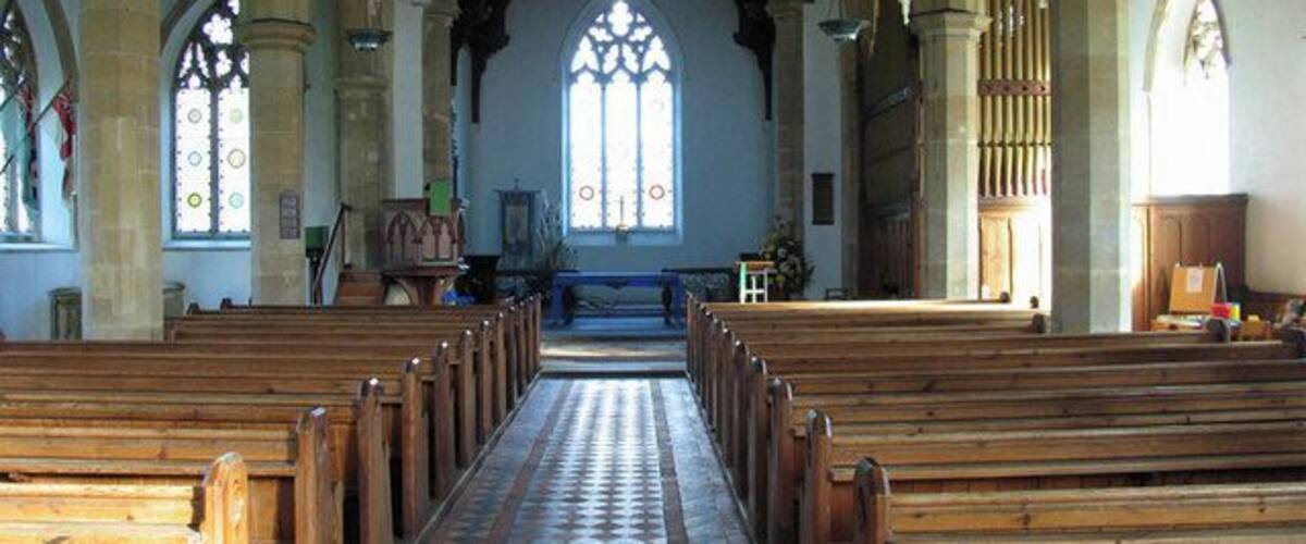 An interior of a church with wooden pews, a tiled floor, and a large stained glass window.