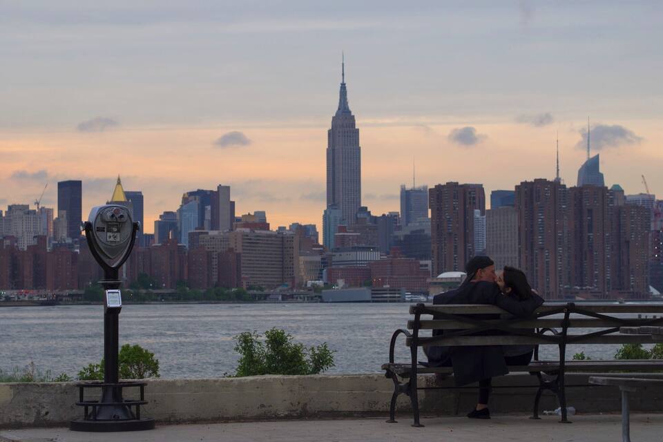 Une vue sur les gratte-ciel d’une ville, avec l’Empire State Building, une rivière et un couple assis sur un banc.