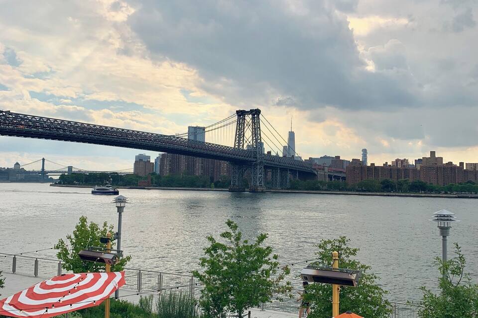 A riverside patio with tables and striped umbrellas, overlooking a bridge and city skyline.