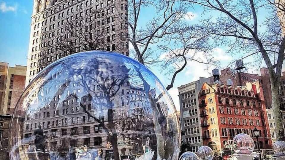 Une grande sphère de verre qui reflète un paysage urbain, avec un immeuble imposant en arrière-plan et un arbre au premier plan.