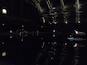 A dimly lit indoor space with a reflective floor, a stage with a few people, and a large industrial structure in the background.