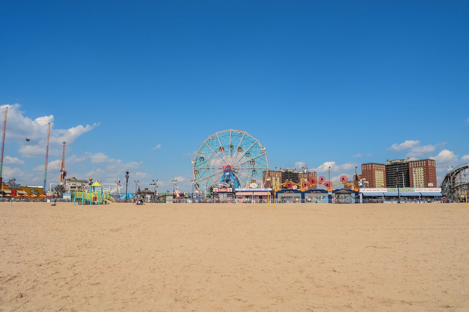 A beach with a large Ferris wheel, amusement rides, and buildings in the background.