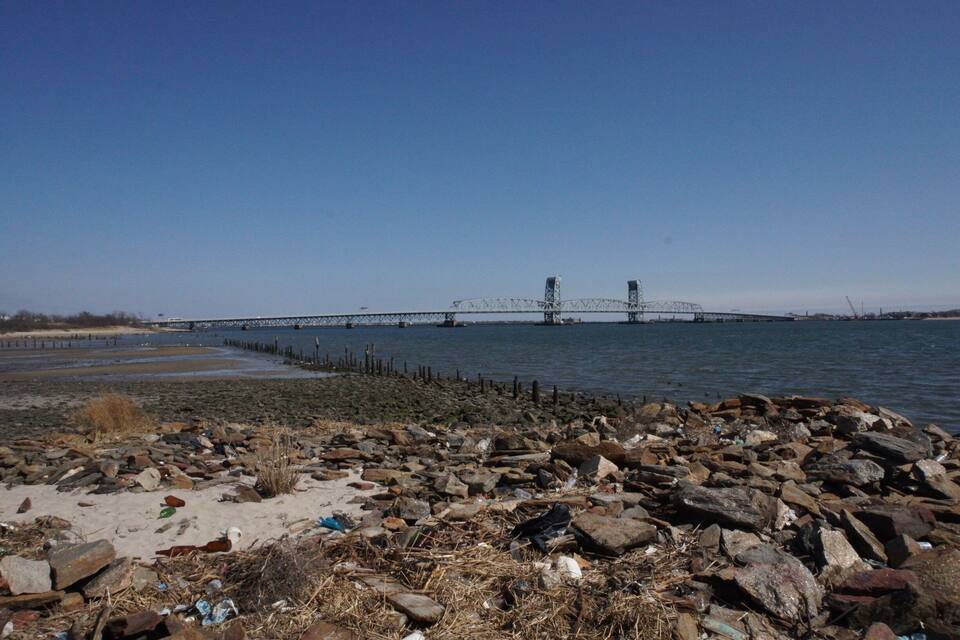 A rocky shoreline with debris and a bridge in the background.