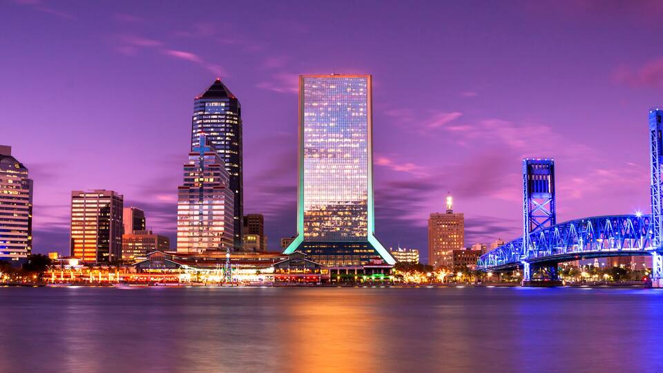 A city skyline with modern skyscrapers and a blue-lit bridge over a body of water.