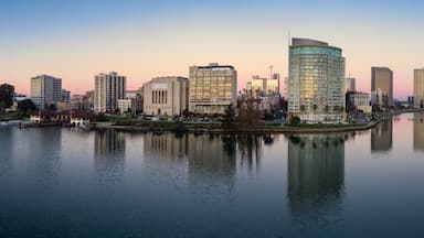 Silhueta urbana com edifícios modernos, um lago tranquilo refletindo as construções, e um céu claro.