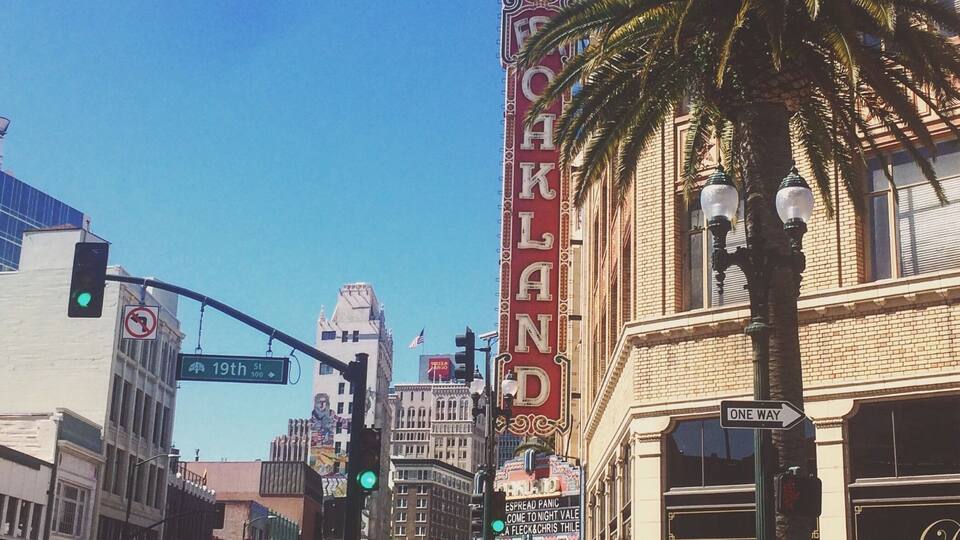 A street corner with a 'Oakland' sign, a '19th St' street sign, and a 'One Way' sign.