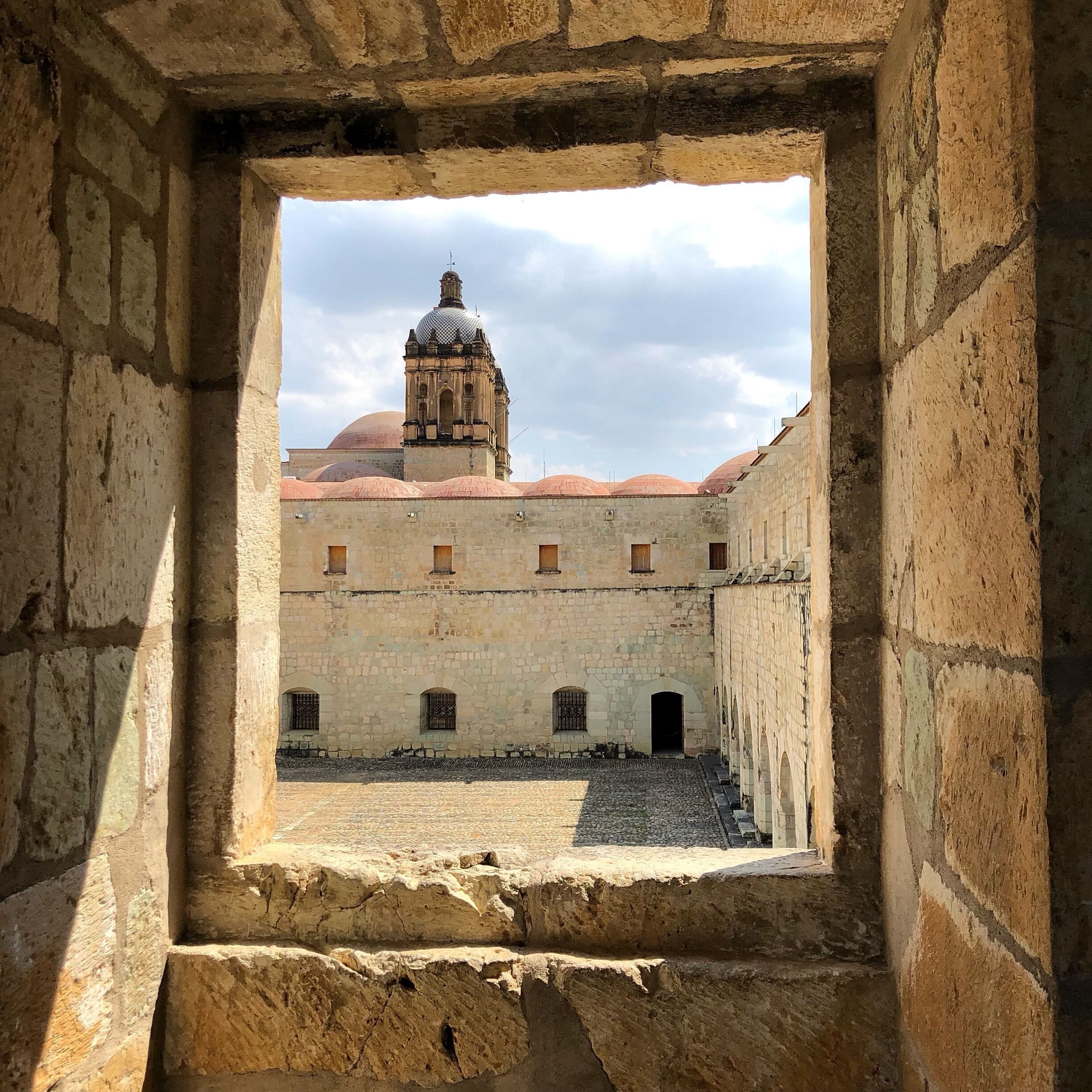 A view through a stone archway of a courtyard with a building featuring a dome and a bell tower.