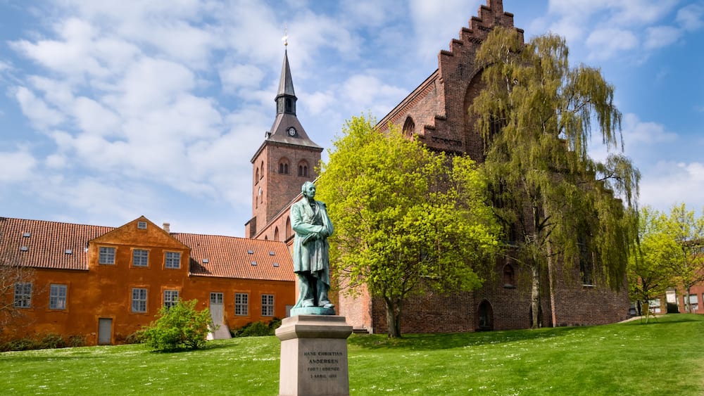 Une statue d’Hans Christian Andersen dans un parc, avec une église et une haute tour en arrière-plan.