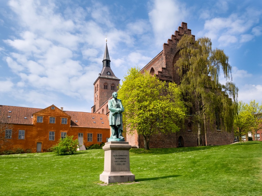 Eine Statue von Hans Christian Andersen in einem Park, im Hintergrund eine Kirche und ein hoher Turm.