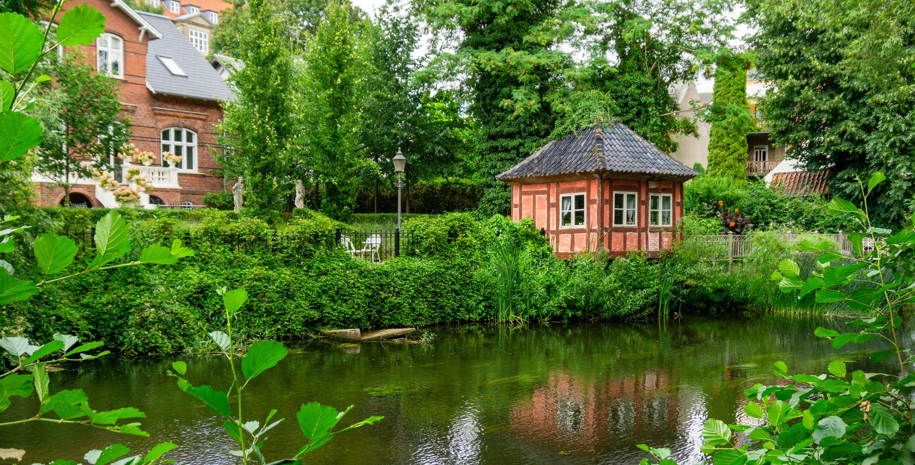 A serene pond with a small red-roofed structure surrounded by lush greenery and a brick house in the background.