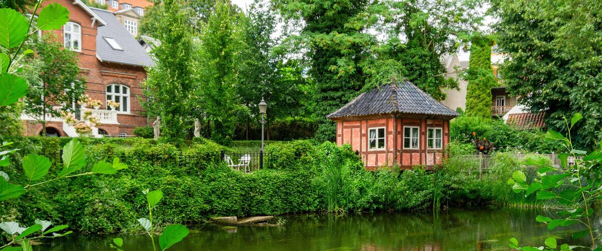 A serene pond with a small red-roofed structure surrounded by lush greenery and a brick house in the background.